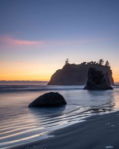 two large rocks sitting on top of a sandy beach next to the ocean at sunset