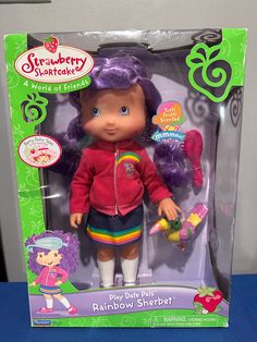 the toy doll is in its box and it has purple hair, wearing a red jacket