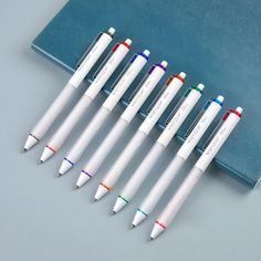 six pens are lined up next to each other on a blue notebook with a book in the background