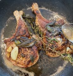 two pieces of meat in a frying pan with some herbs on the side and a spoon