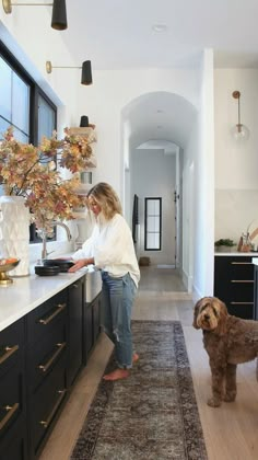 a woman standing in a kitchen next to a dog on a rug and looking at the floor