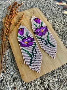 the beaded earrings are decorated with purple flowers and green leaves on a wooden board