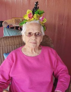 an elderly woman wearing a flower crown on her head sitting in a wicker chair