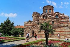 people are walking around in front of an old brick building with many flowers and trees