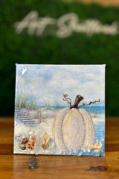 a card with a painting of a pumpkin and seashells in the sand on a wooden table