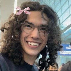 a woman with curly hair wearing glasses and a pink bow on her head smiles at the camera