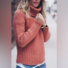 a woman in an orange sweater and jeans is standing on the street with her hand under her chin