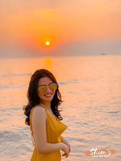 a woman in a yellow dress standing on the beach at sunset with her arms around her waist