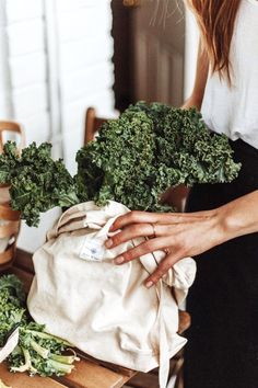 a woman standing next to a bag filled with green leafy vegetables on top of a wooden table