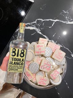 a person holding a bottle of alcohol next to some cookies on a plate with writing on them