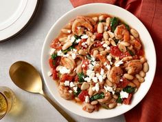 a white bowl filled with pasta and shrimp on top of a red table cloth next to two gold spoons