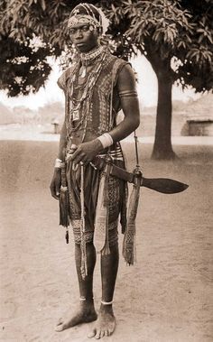 an old photo of a man in native clothing holding two paddles and standing under a tree