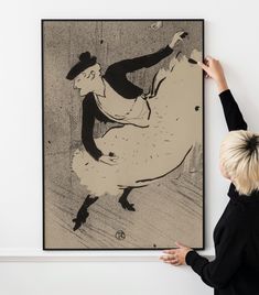 a woman is holding up a painting in front of a wall with a black and white drawing on it