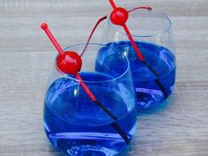 two glasses filled with blue liquid and cherries on the rims, sitting on a table