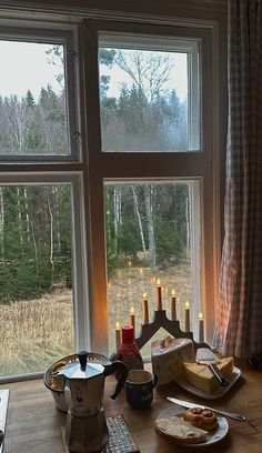 a table with food and candles on it in front of a window that looks out onto the woods