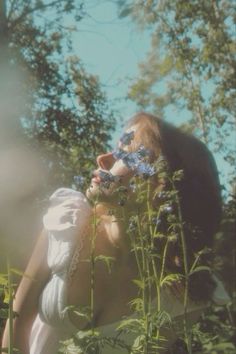 a woman in white dress smelling flowers with trees in the background behind her