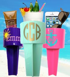 three different colored cups on the beach with cell phones and sunscreens in them