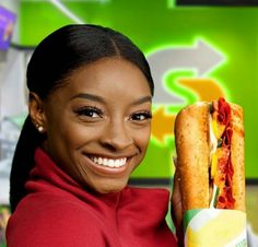 a smiling woman holding a sub sandwich in front of her face and looking at the camera