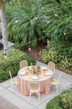 an outdoor dining table set up with pink linens and white chairs, surrounded by greenery