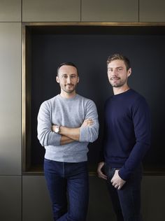 two men standing next to each other in front of a black and silver wall with their arms crossed