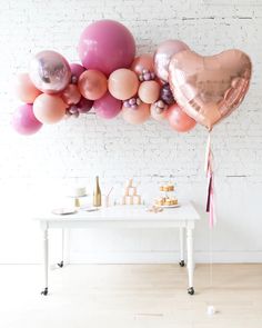 a white table topped with lots of pink and gold balloons next to a brick wall