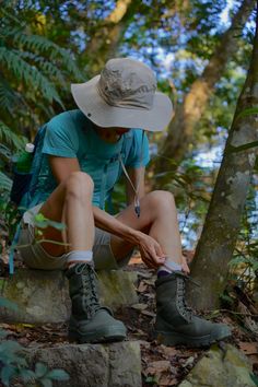 a man sitting on top of a rock next to a tree wearing a hat and hiking boots