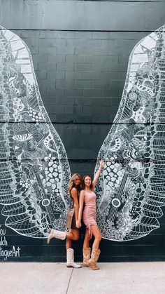 two women standing in front of a wall with wings painted on it