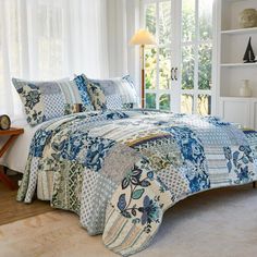 a bed in a room with white walls and curtains on the window sill is covered by a blue patchwork quilt