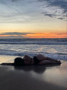 a woman laying on top of a sandy beach next to the ocean at sunset or dawn