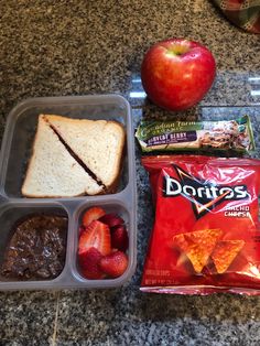 lunch box with sandwich, fruit and chips on granite counter top next to an apple