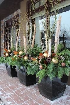 three black planters filled with evergreen, pine cones and gold baubles on a brick patio