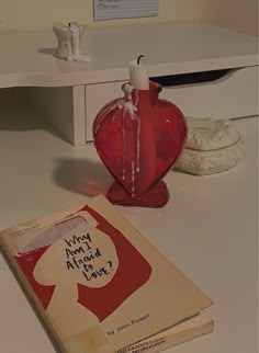 a red heart shaped vase sitting on top of a table next to an open book