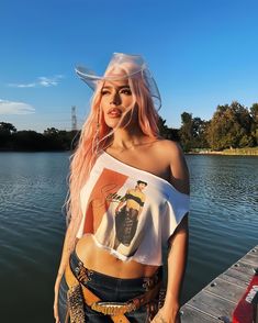 a woman with pink hair standing on a dock next to the water wearing a cowboy hat