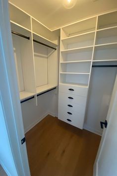 an empty walk in closet with white shelves and drawers on the wall, next to a wooden floor