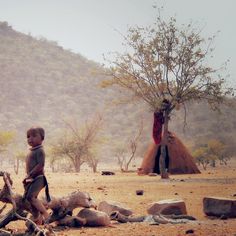 a young boy standing next to a tree on top of a dirt field with mountains in the background