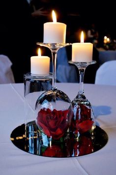 three wine glasses with red roses in them on a table next to two white candles