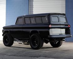 an old black truck parked in front of a blue building with large tires on it