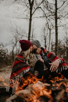 a man and woman sitting next to each other in front of a campfire with their arms around each other