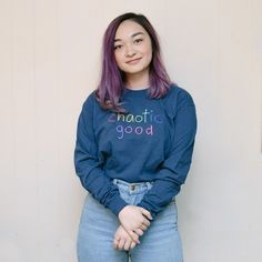 a woman with purple hair wearing a blue sweatshirt and jeans standing in front of a white wall