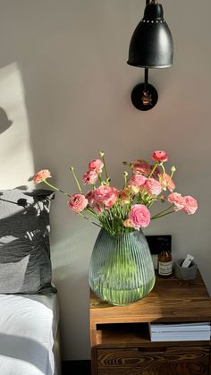 a green vase filled with pink flowers sitting on top of a wooden table next to a bed