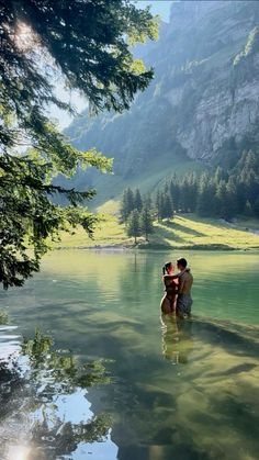 two people are sitting on the edge of a body of water with mountains in the background