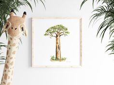 a giraffe standing next to a tree in front of a white wall with a painting on it