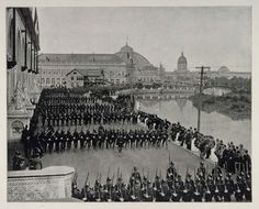 an old black and white photo of men in uniforms marching down a street with buildings in the background