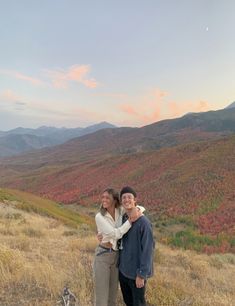 a man and woman standing on top of a grass covered hillside with mountains in the background