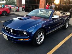 a blue mustang convertible parked in front of a brick building with people standing around it