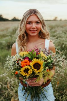 a woman standing in a field holding a bouquet of sunflowers and greenery