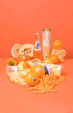 oranges and other food items on an orange colored surface with two cans of soda in the background