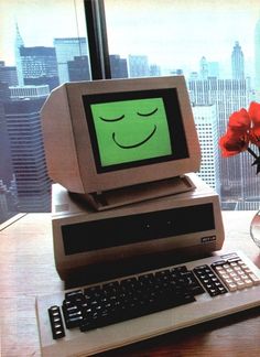 an old computer with a smiley face on the screen sitting next to a flower in a vase