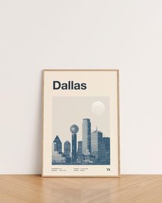 a framed poster with the word dallas in front of a white wall and wooden table