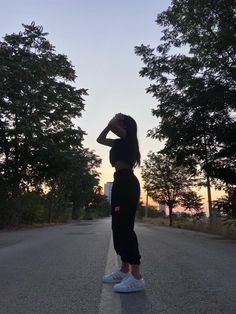 a woman standing on the side of a road with her hands behind her head and trees in the background